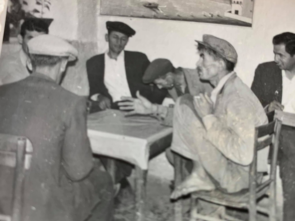 Old black-and-white photograph of village men in caps talking around a table in the coffeehouse